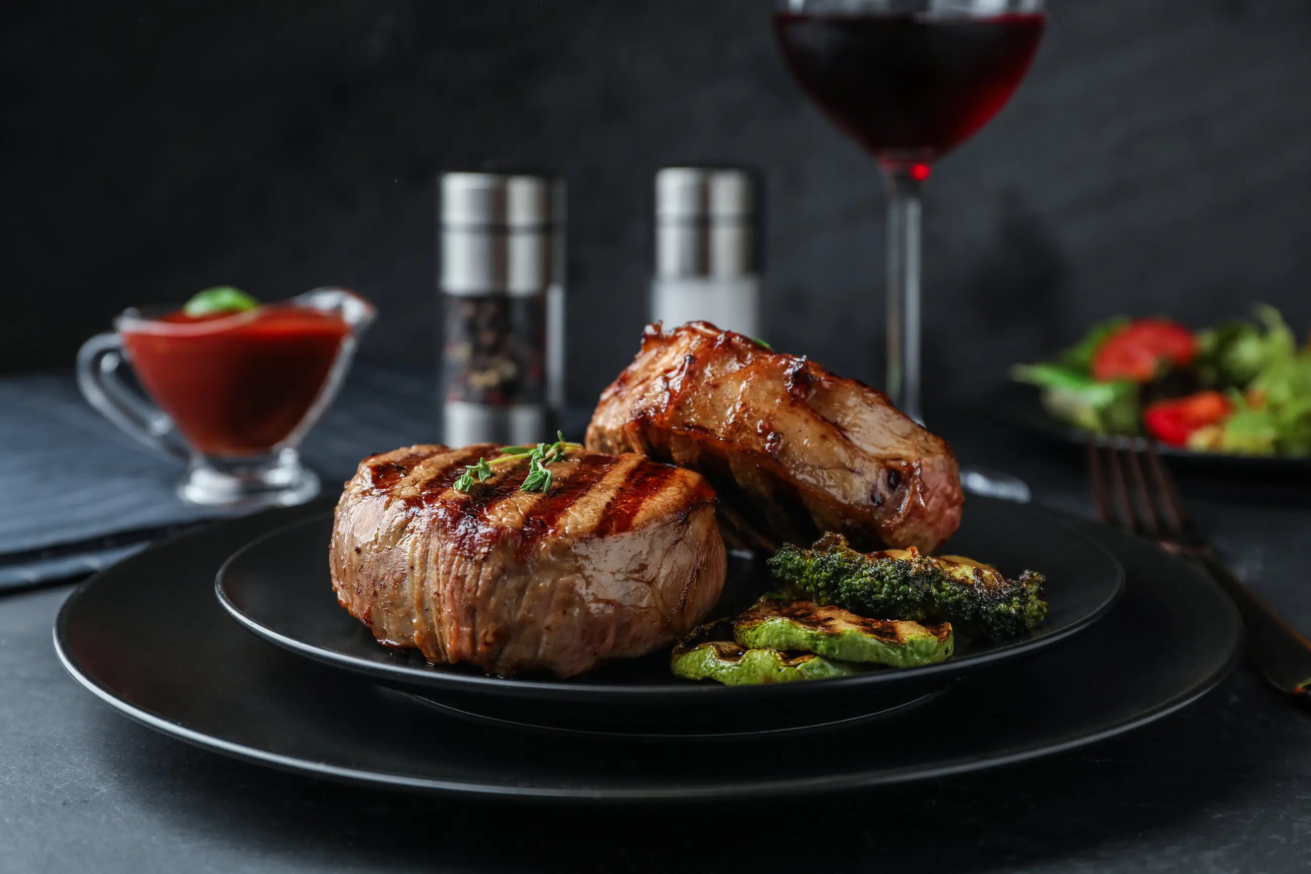 Gegrillte Steaks mit Brokkoli und Paprika auf schwarzem Teller, dahinter ein Glas Rotwein